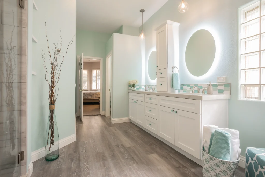 Modern aqua bathroom with white cabinetry, glass shower, and warm wood-like flooring.