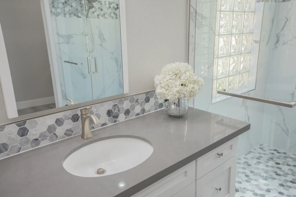 Elegant bathroom with a light stone countertop, gold faucet, hexagonal tiles, and frosted glass shower.