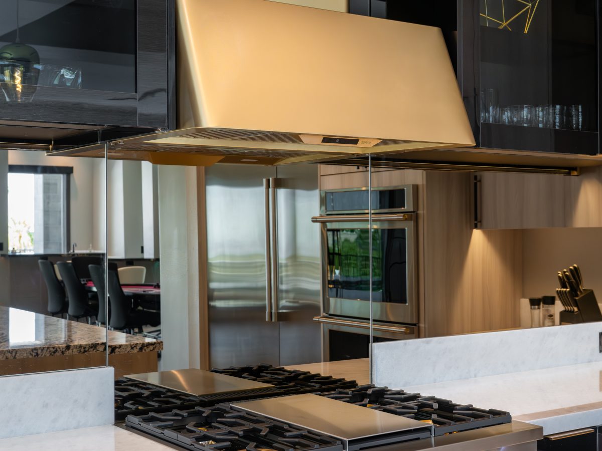 Luxury kitchen with gold range hood, mirrored backsplash, and dark charcoal glossy cabinetry, Reveal Interior Design Las Vegas