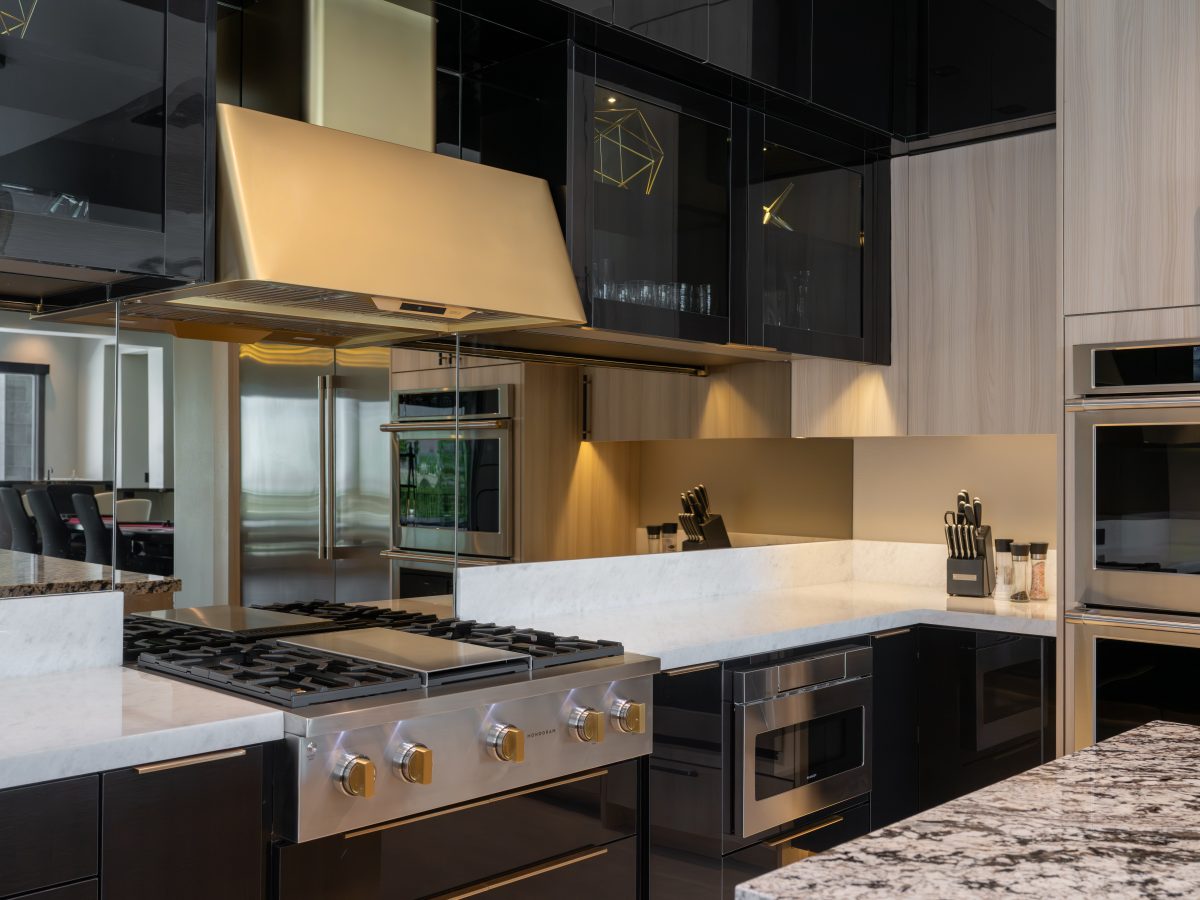 Luxury kitchen with gold range hood, mirrored backsplash, and dark charcoal glossy cabinetry, Reveal Interior Design Las Vegas