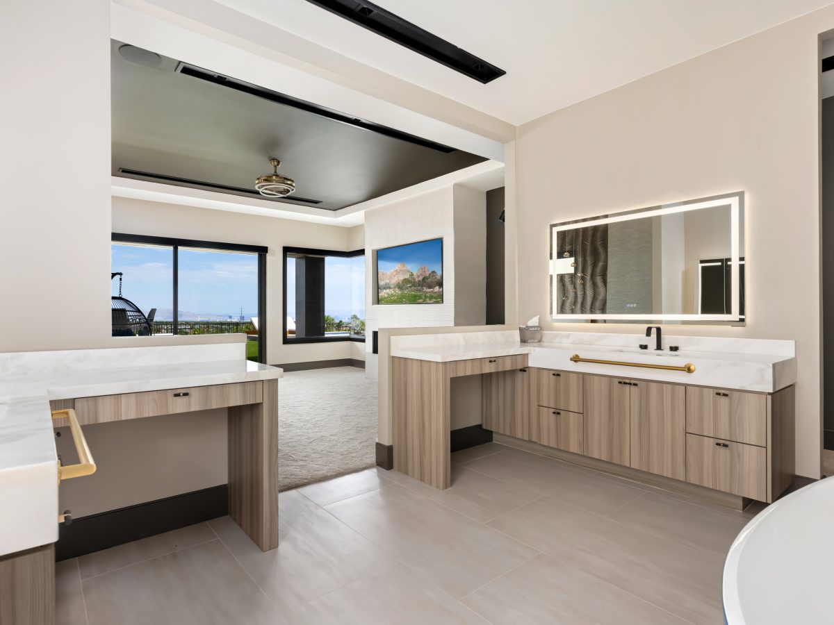 Primary bathroom with warm oak-look vanities, marble-look countertops, and LED mirrors opening to bedroom views, Reveal Interior Design Las Vegas