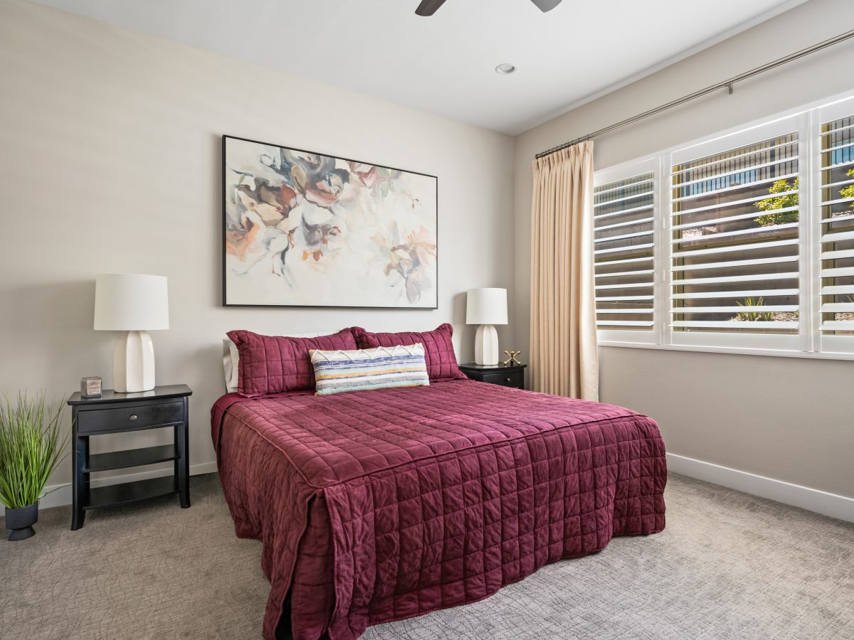 Primary bedroom with deep burgundy quilted bedding, plantation shutters, and floral artwork, Reveal Interior Design Las Vegas