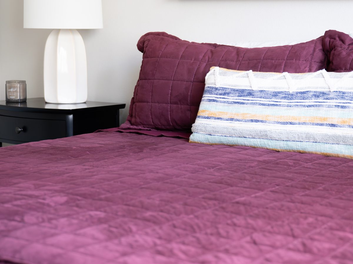 Primary bedroom with plum quilted bedding, striped accent pillow, and abstract artwork, Reveal Interior Design Las Vegas