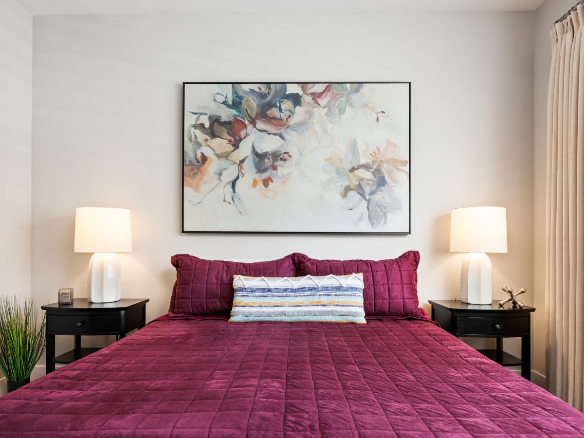 Primary bedroom with cranberry quilted comforter, large contemporary floral artwork, and sand-tone draperies, Reveal Interior Design Las Vegas