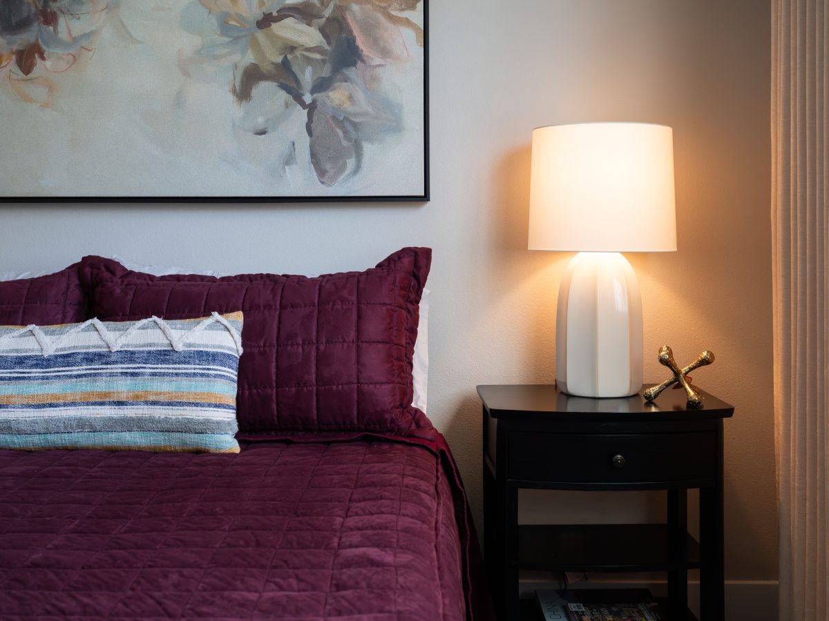 Primary bedroom with cranberry quilted bedding, contemporary floral art, and glowing nightstand lamp, Reveal Interior Design Las Vegas