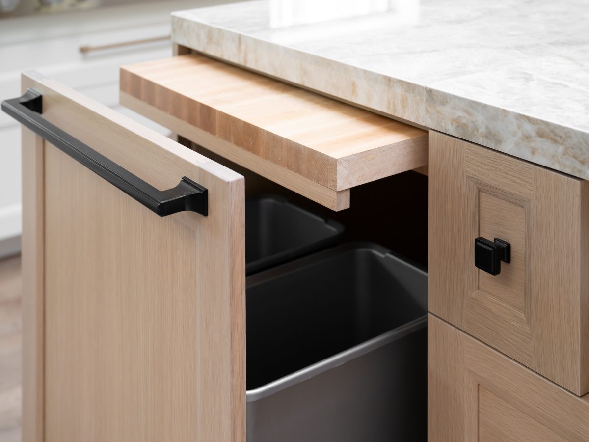 Kitchen island trash pullout with hidden cutting board drawer and light oak cabinetry, Reveal Interior Design Las Vegas