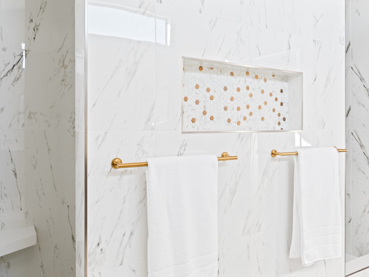 Primary bathroom shower wall with Calacatta-look porcelain tile, gold hexagon mosaic niche, and brushed gold towel bars, Reveal Interior Design Las Vegas