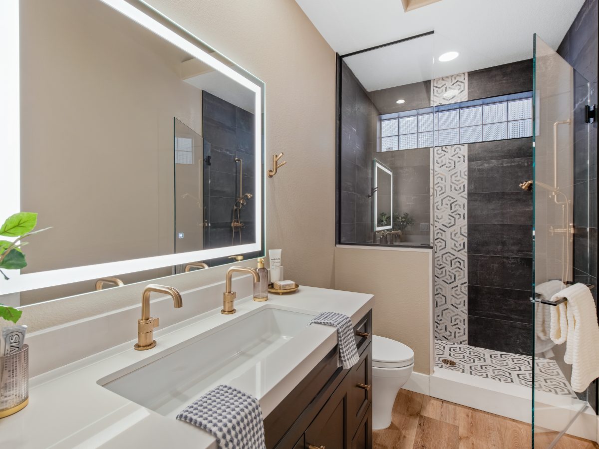 Modern guest bathroom with brushed gold fixtures, backlit mirror, and geometric tile shower, Reveal Interior Design Las Vegas